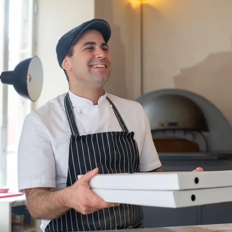 A pizzeria owner in a striped apron carrying boxed orders past the oven